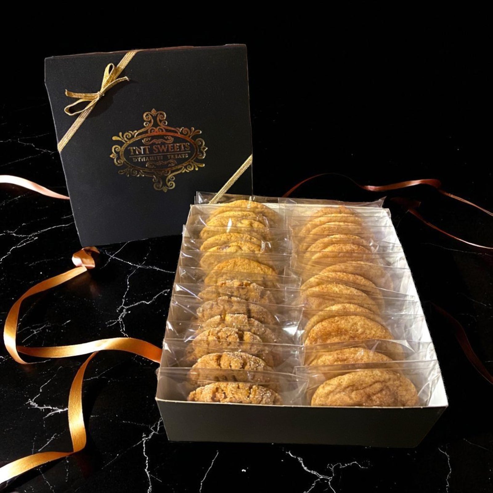 Gift box of cookies with a glossy black box featuring gold ribbon on a black marble surface.