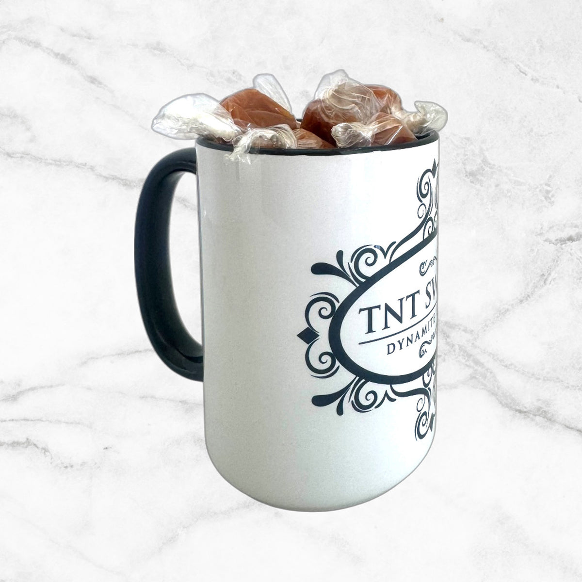 15oz TNT Signature White Mug with Caramels unwrapped with a black handle on the side on a white marble background.