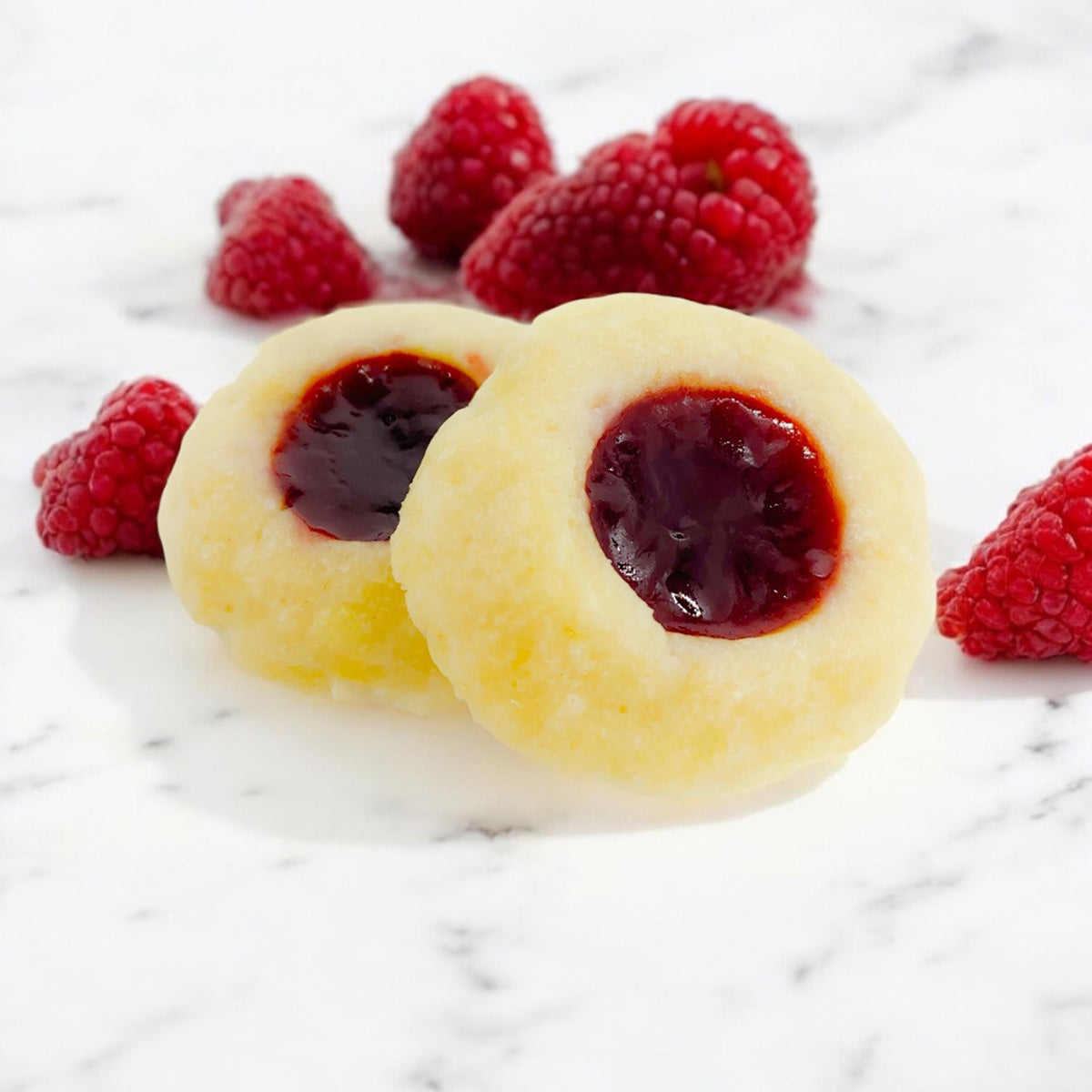 Two Raspberry Thumbprint cookies on a marble countertop with scattered raspberries in the background.