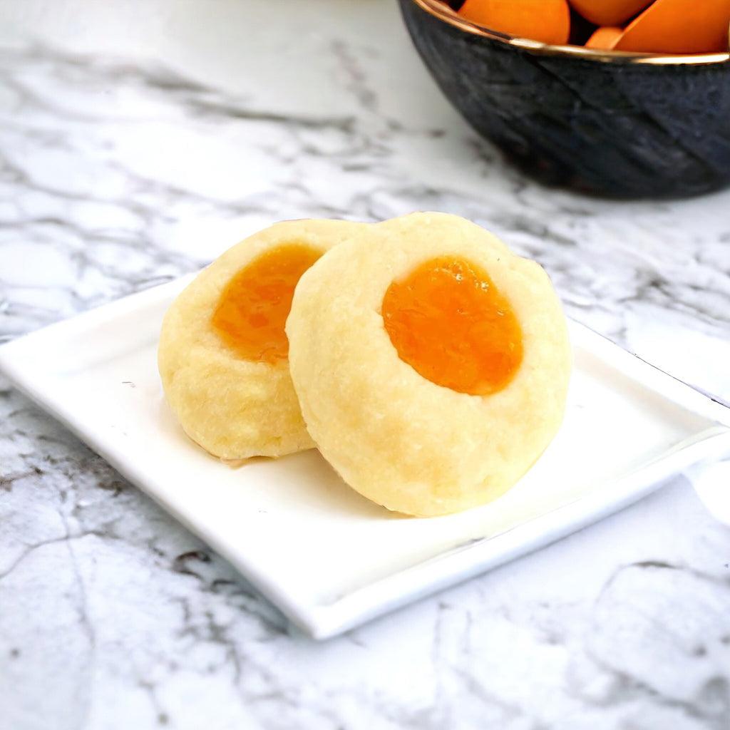 Two apricot thumbprint cookies on a plate on a marble countertop with a bowl of apricots in the background. 