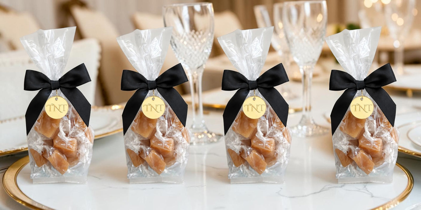 Four clear cellophane gift bags filled with Amber Velvet sea salt caramels, tied with black ribbon bows and hanging TNT gold medallion arranged on a white marble table with wine glasses in a softly lit indoor setting.