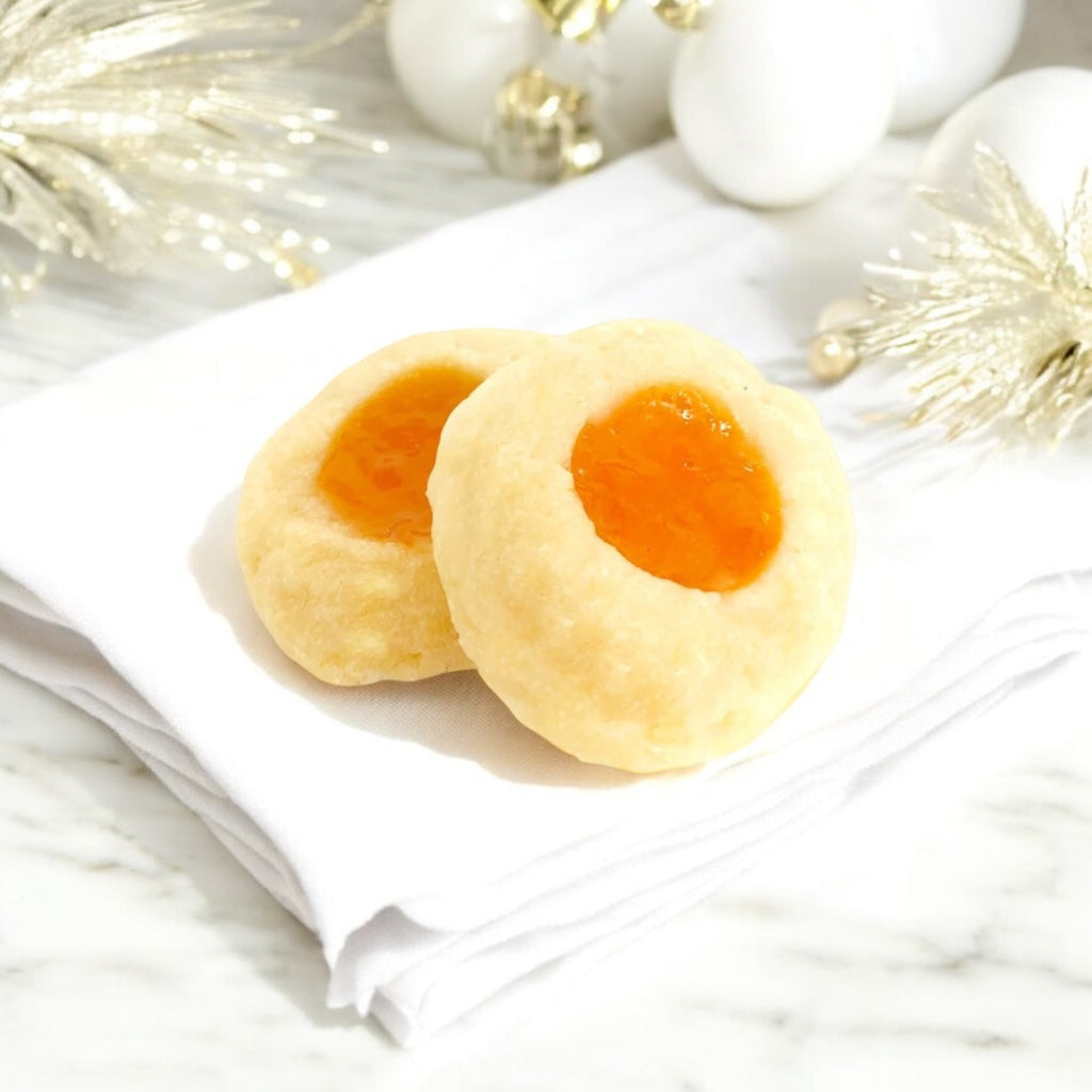 Two Apricot Thumbprint cookies on a linen napkin on a white marble countertop with white and gold decor in the background.