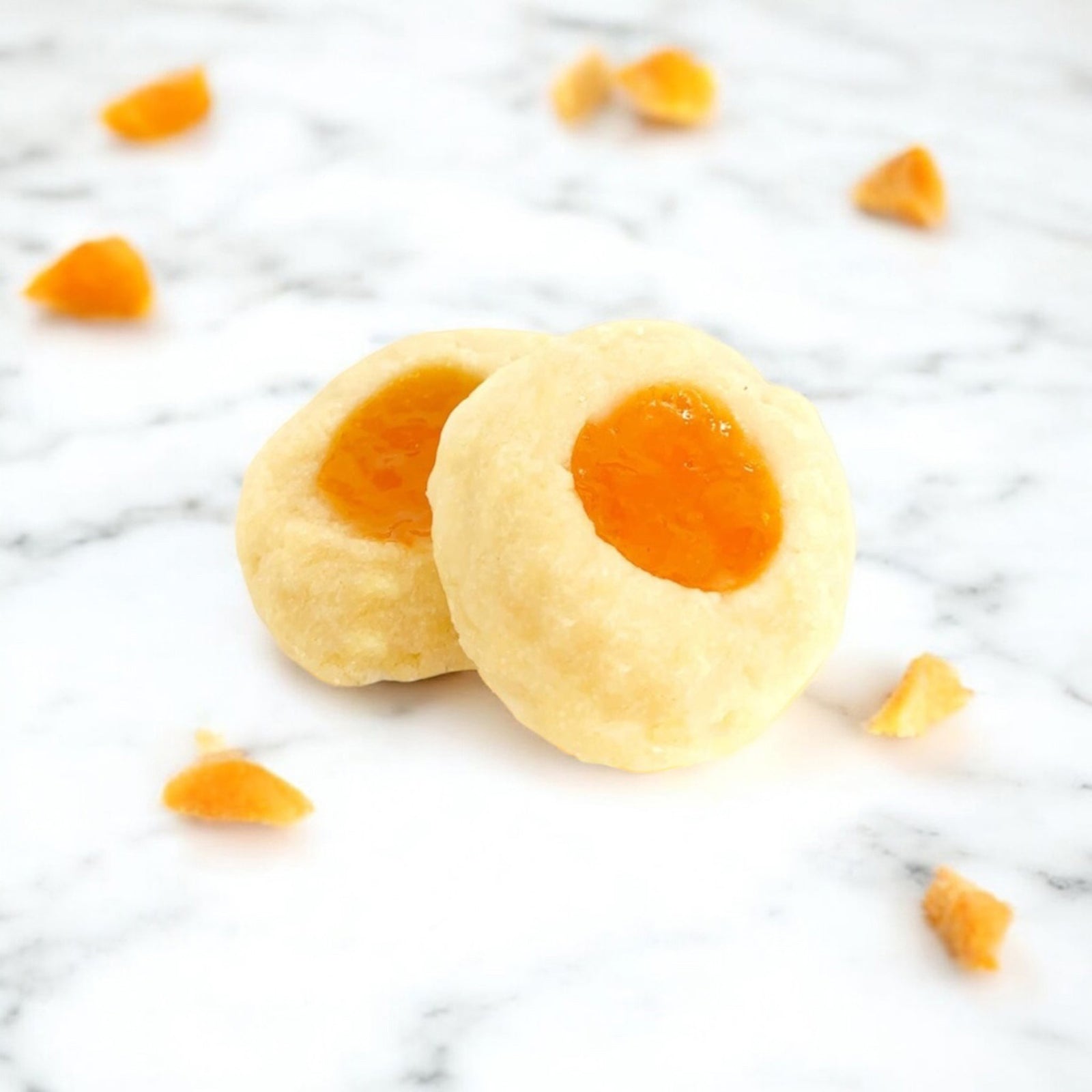 Two Apricot Thumbprint cookies on a white marble countertop with scattered apricots.