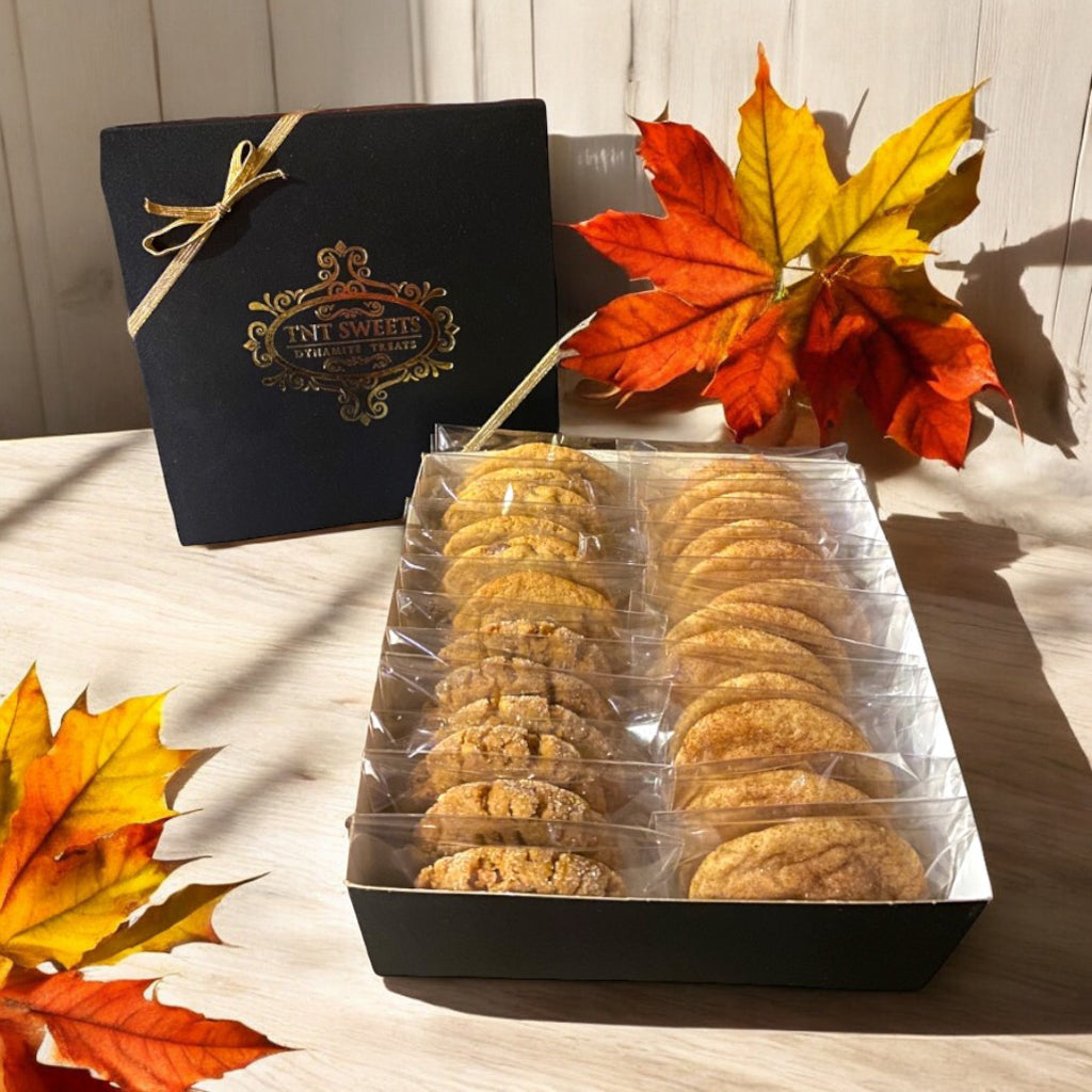 An assortment of cookies in a TNT Sweets black signature cookie box on a wooden countertop with scattered fall leaves.