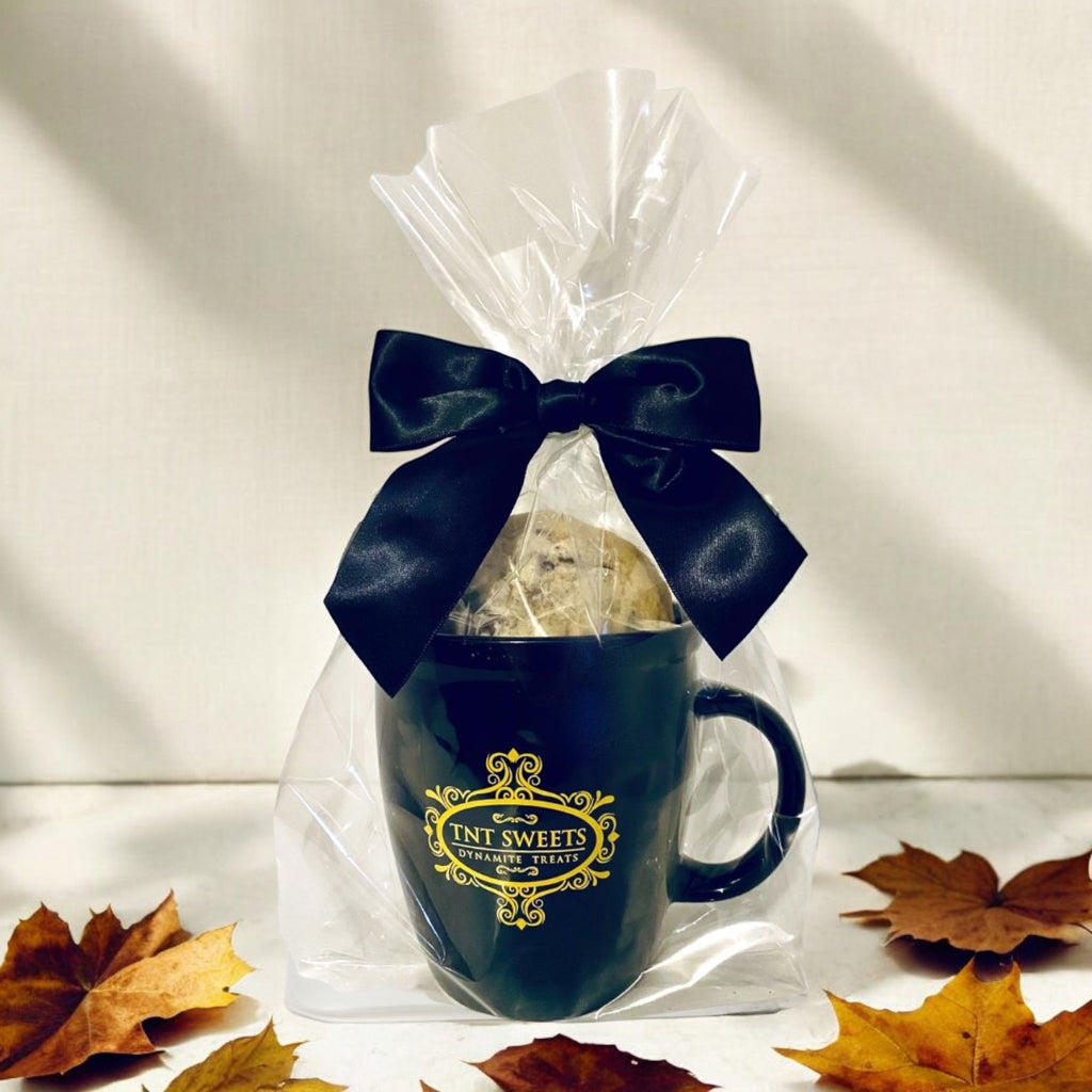 Chocolate Chip cookies filled in an 18oz mug wrapped in cellophane with a black bow on a wooden countertop with scattered fall leaves.