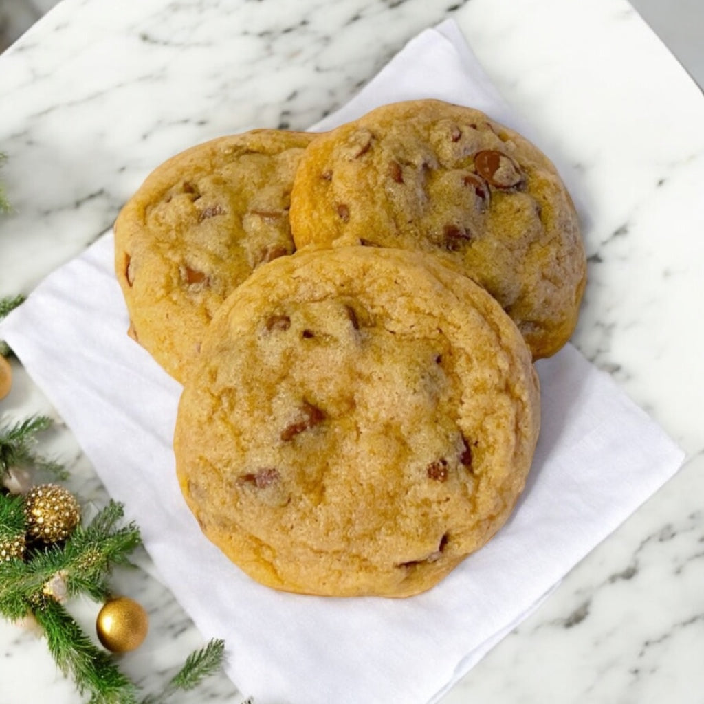 Three chocolate chip cookies on a linen napkin on a marble table with pine and gold holiday decor. 