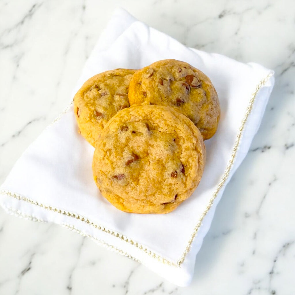 Three chocolate chip cookies on a linen napkin with gold trim on a marble countertop.