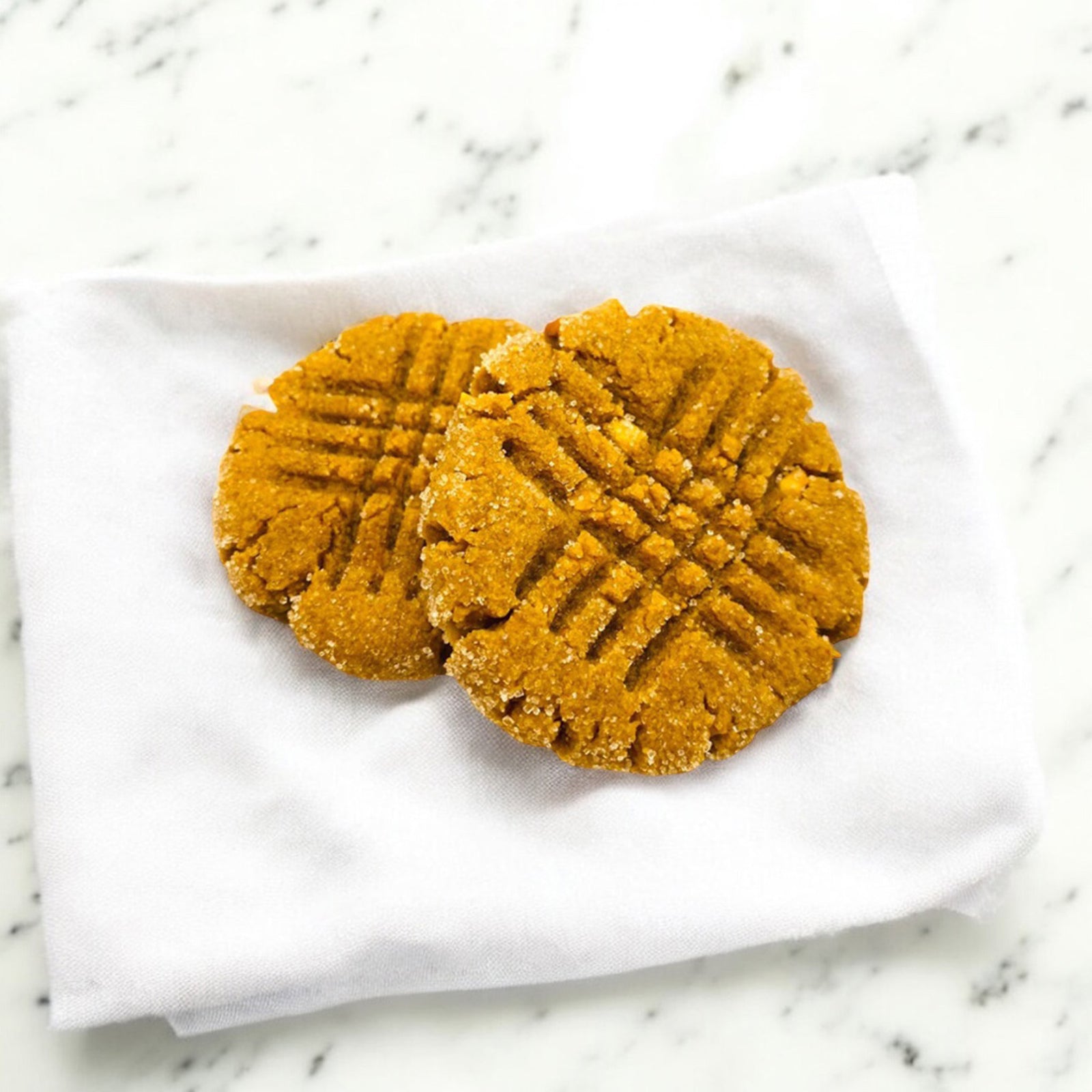 Two classic peanut butter cookies on a white linen napkin on a white marble countertop.