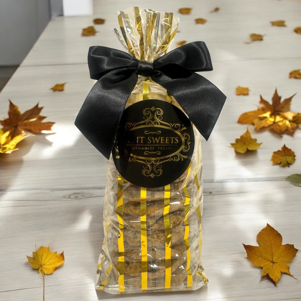 An assortment of cookies in a TNT Sweets gold striped gift bag on a wooden countertop with scattered fall leaves.