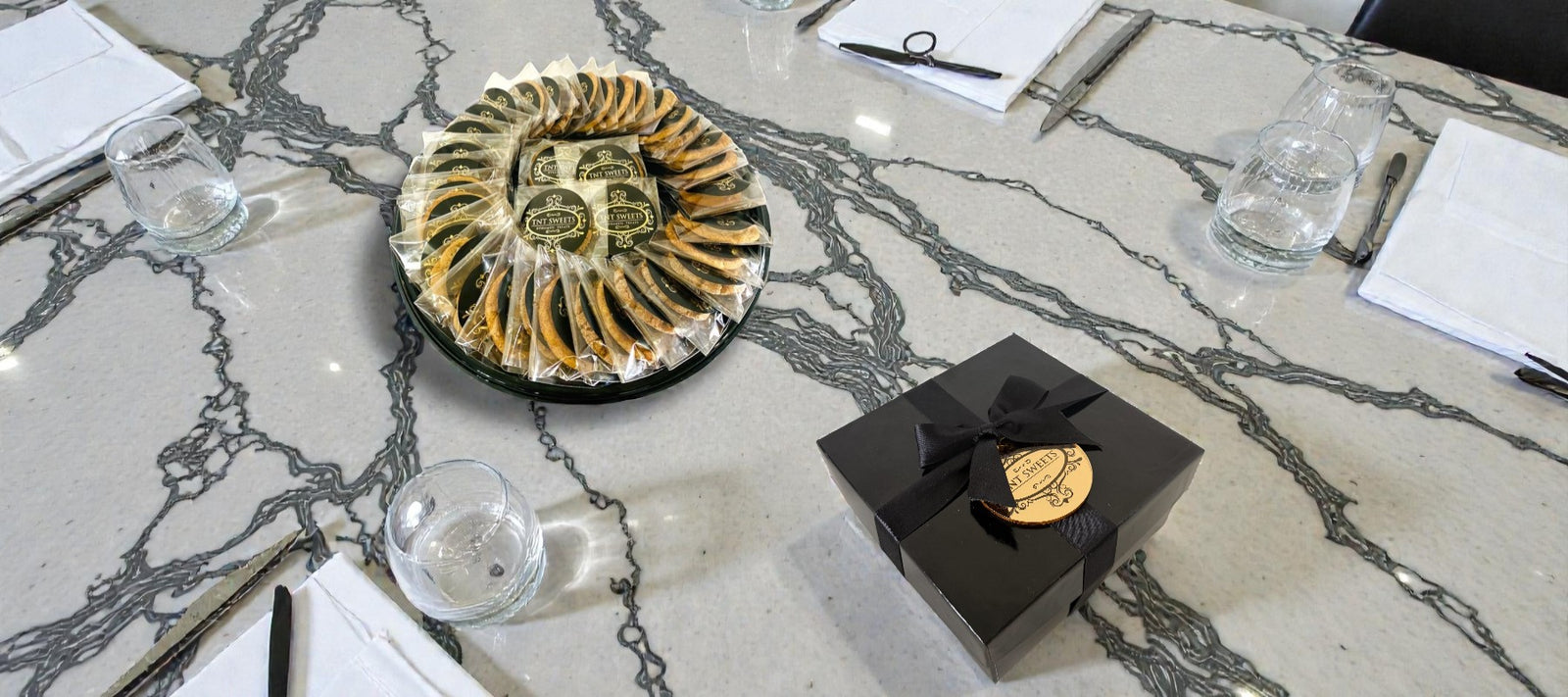 Corporate executive table setting with a black box, glassware, and a cookie platter and black cookie box with a gold medallion on a marble surface.
