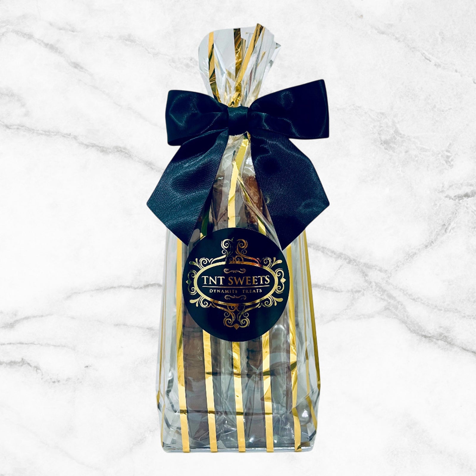 Twelve dark chocolate pretzels in a gold cello bag with a black satin bow and TNT Sweets logo on a white marble background.