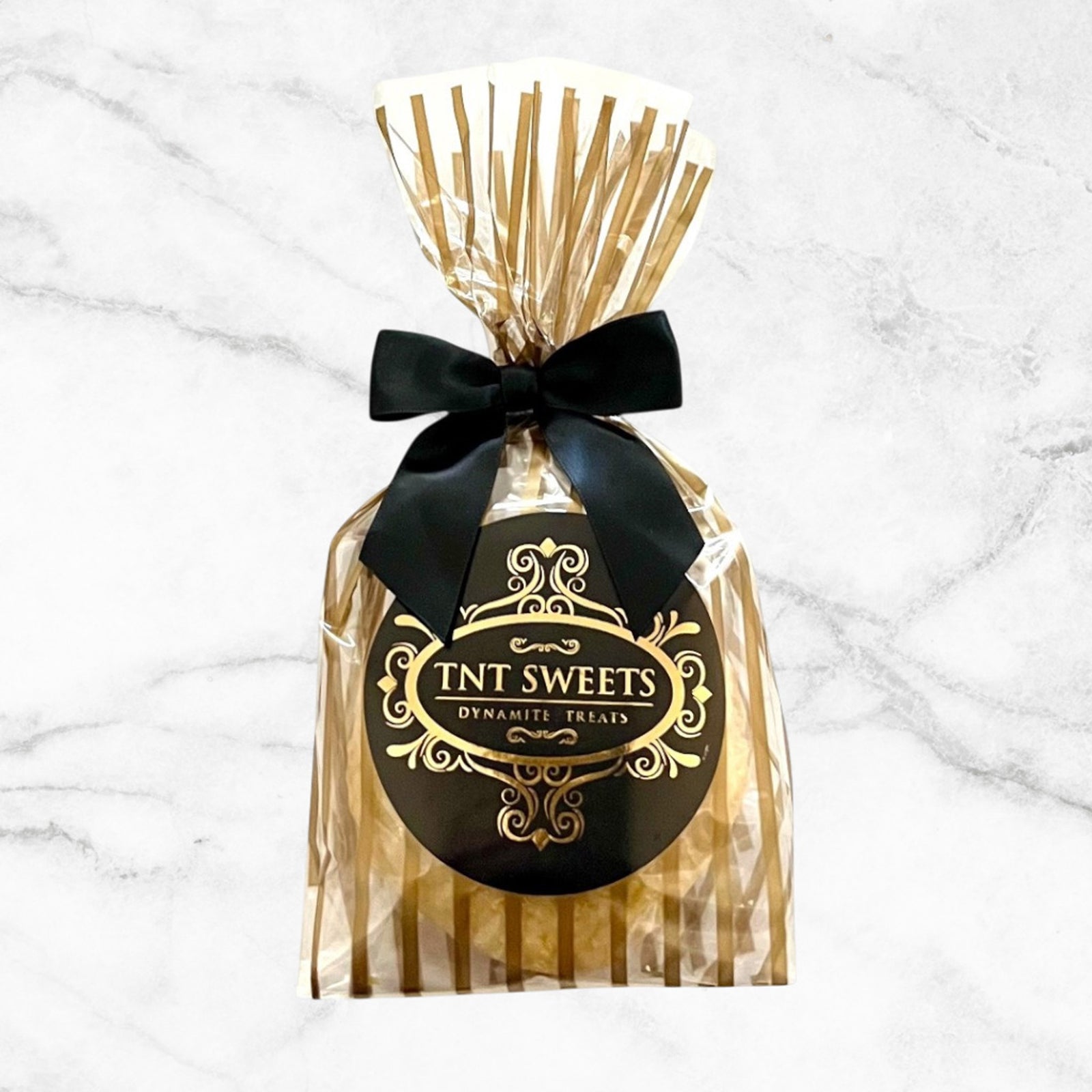 Two cookies in a gold striped bag with black bow and logo on a white marble background. 