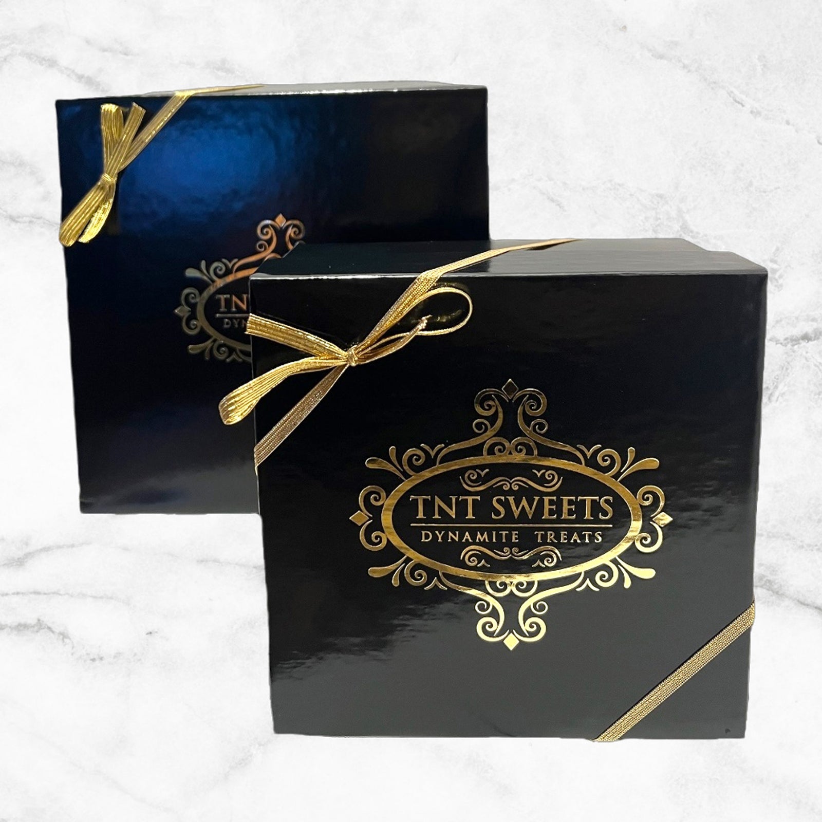 Two black gift boxes with gold logos and gold ribbons on a white marble background.
