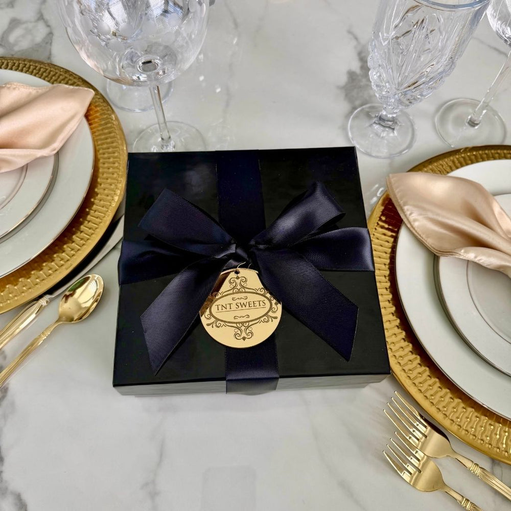 TNT Sweets Keepsake Box with black satin ribbon and gold acryclic medallion on a marble dining table with gold place settings and crystal.