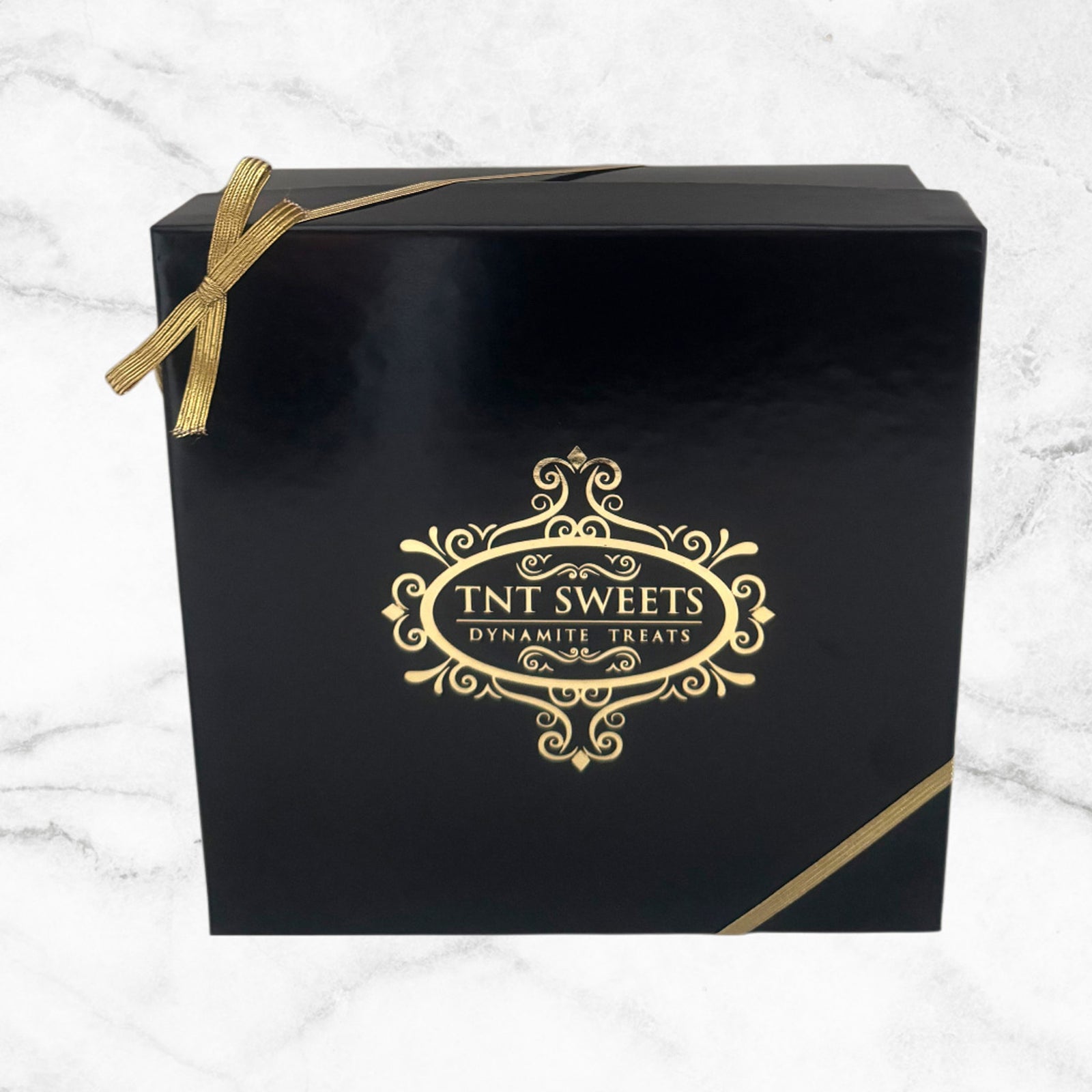 Black box with gold bow and TNT Sweets logo on a white marble background.