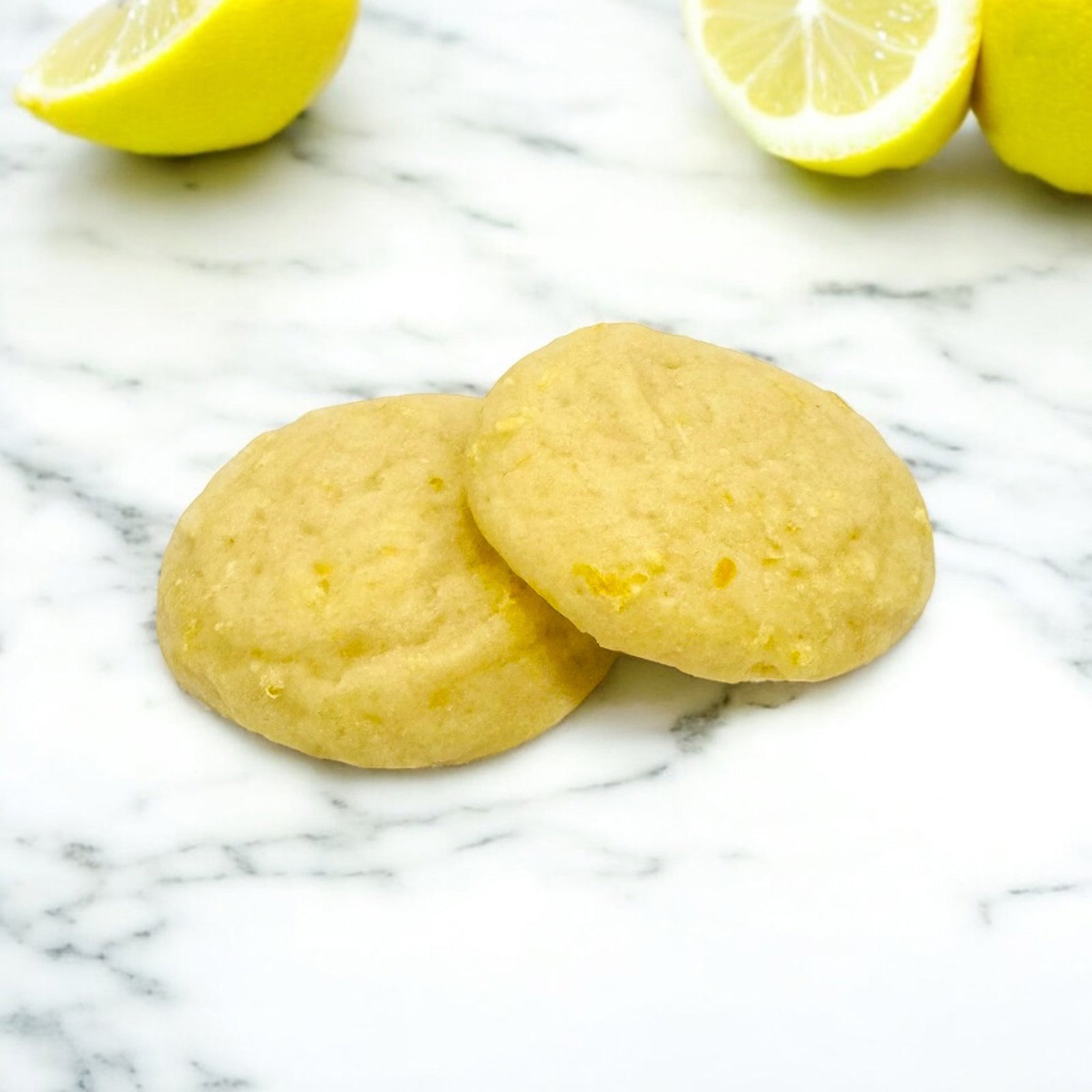 Two lemon cookies on a marble surface with lemon halves in the background.