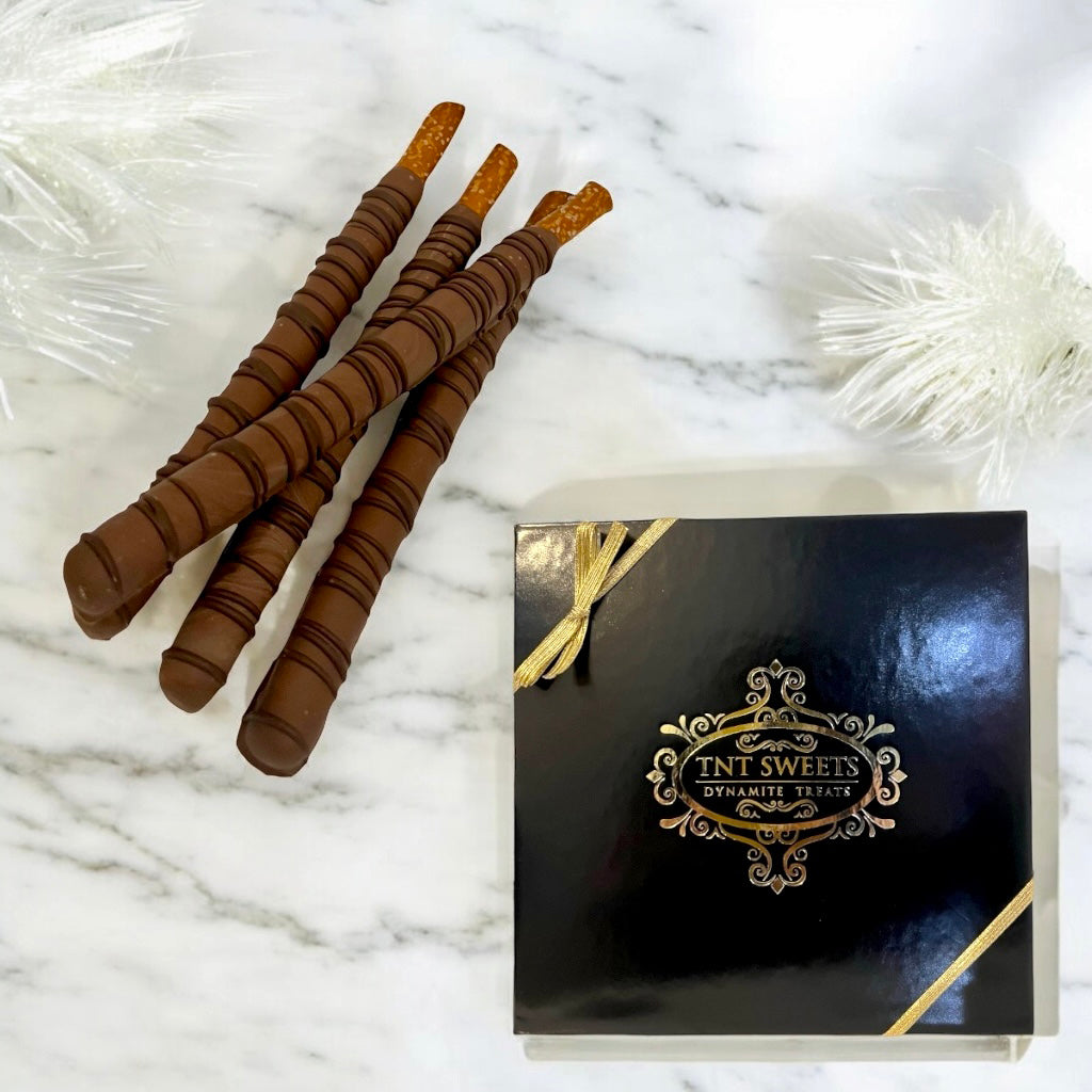 Stacked milk chocolate pretzels on a marble countertop with a TNT Sweets black gift box displayed and scattered white decor.