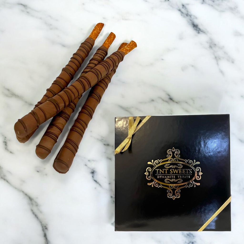 Milk chocolate dipped pretzels next to a black and gold gift box on a white marble countertop.