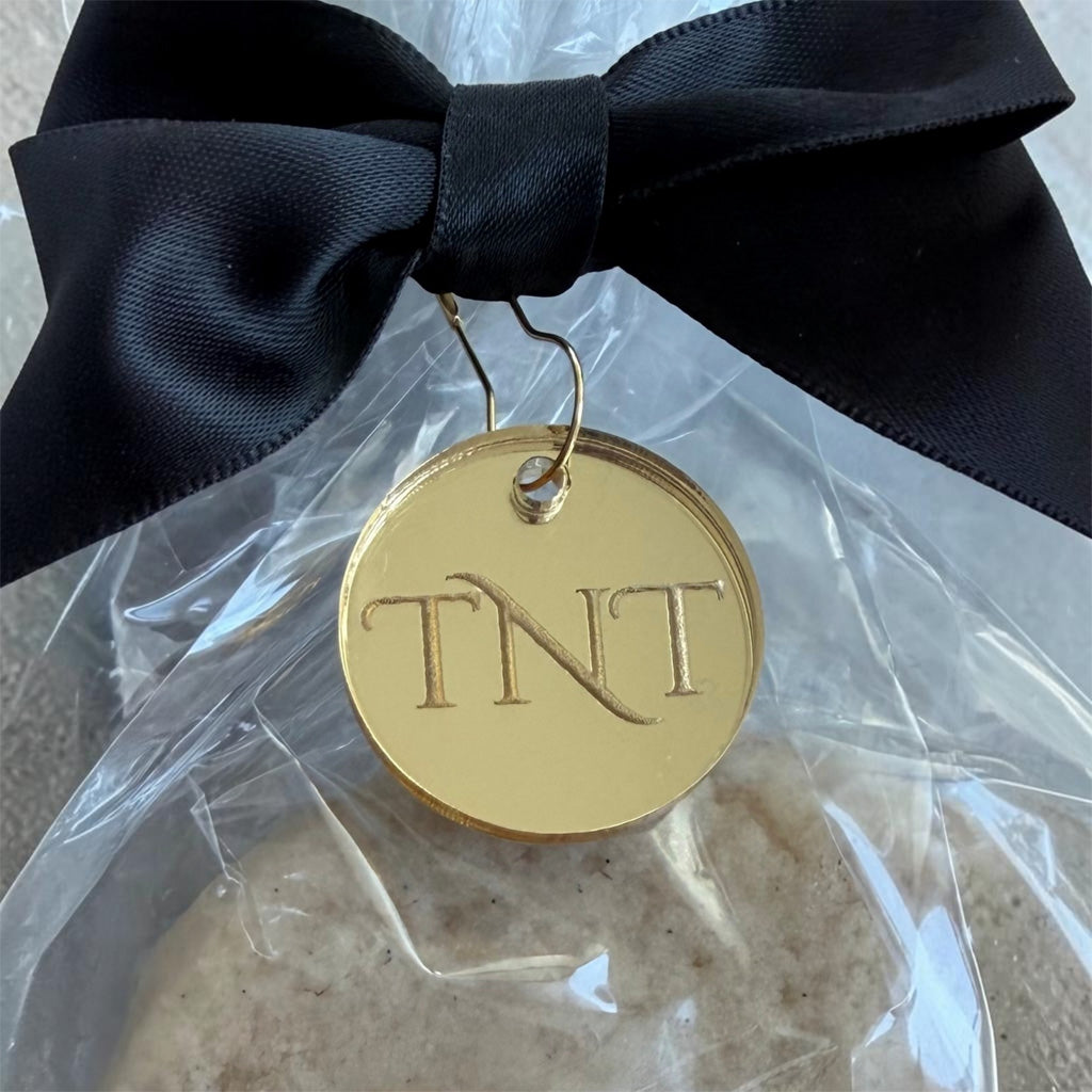 Close up of TNT gold acrylic monogram affixed to a black ribbon and partial view of The Premiere Vanilla Bean Shortbread cookie.