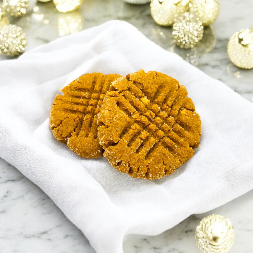 Two peanut butter cookies on a linen napkin with on a marble countertop with scattered white decor in the background. 