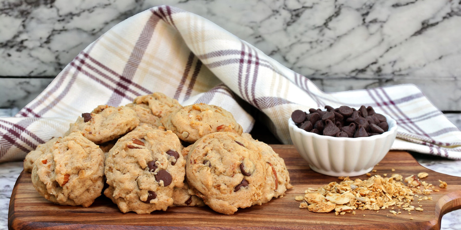 Pecan Chocolate Chip cookies on a wood block with a small bowl of chocolate chips and pecan scatter with a linen napkin on a marble countertop.