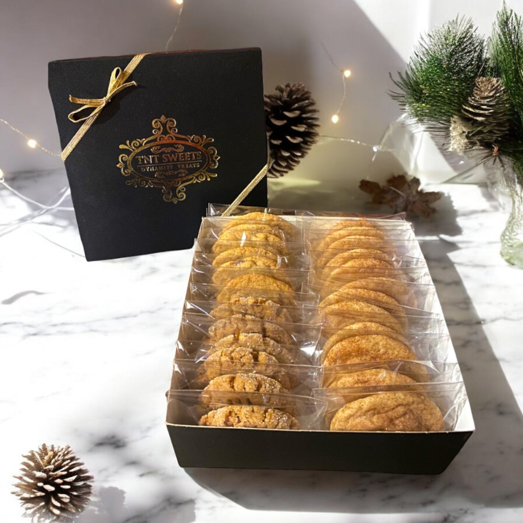 An assortment of cookies in a TNT Sweets black signature cookie box on a marble countertop with holiday decor.