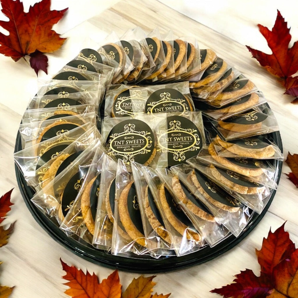 An assortment of three dozen cookies on a black cookie platter on a wooden countertop with scattered fall leaves.