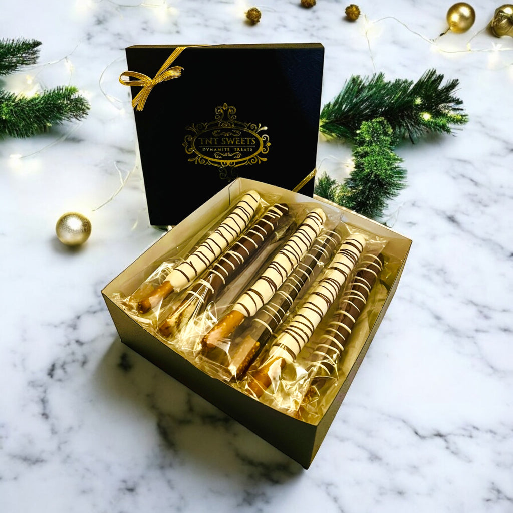 White chocolate, milk chocolate and dark chocolate pretzels in the black and gold signature gift box on a  marble countertop with a holiday background.