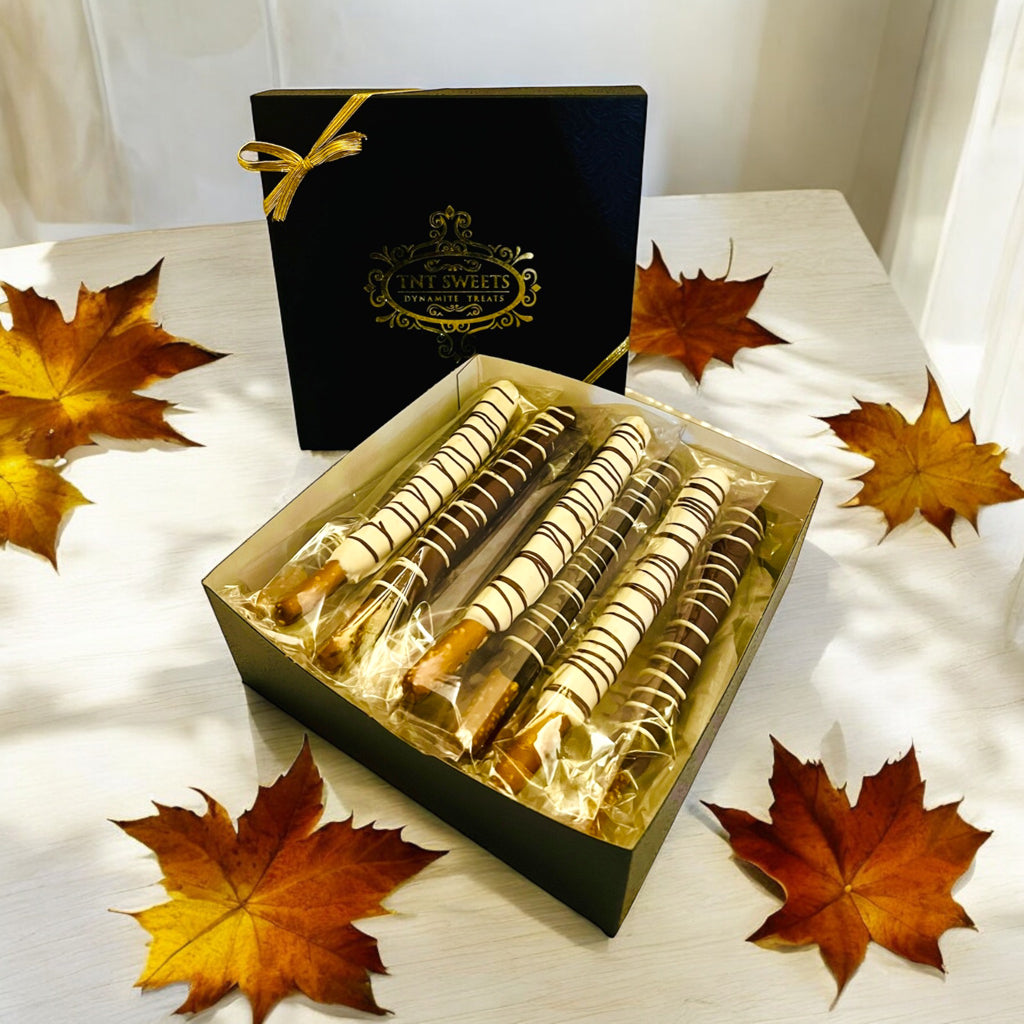 White chocolate, milk chocolate and dark chocolate pretzels in our  signature black box on a wooden countertop with scattered fall leaves in the background.