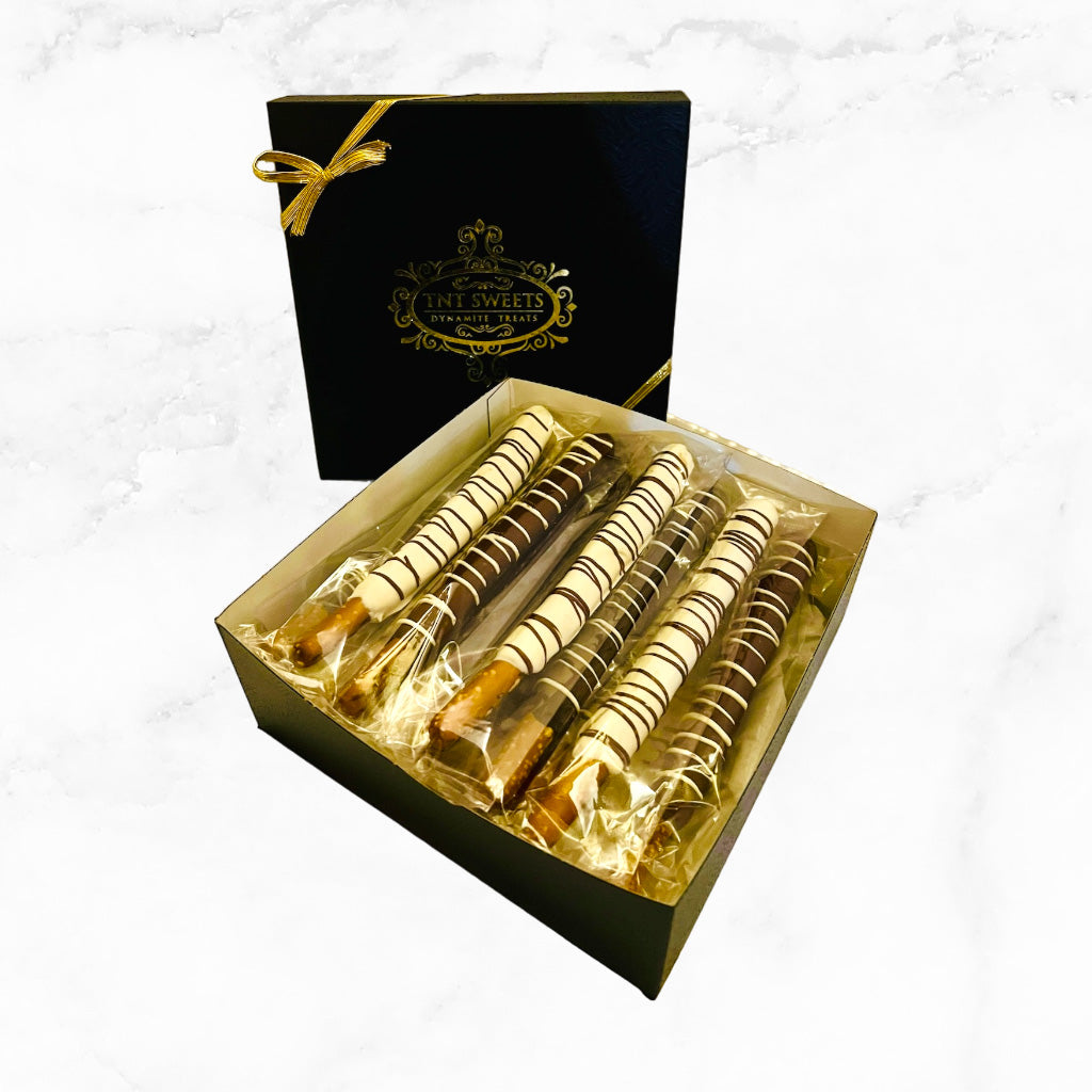White chocolate, milk chocolate and dark chocolate pretzels in the black and gold signature gift box on a white marble countertop with a holiday background.