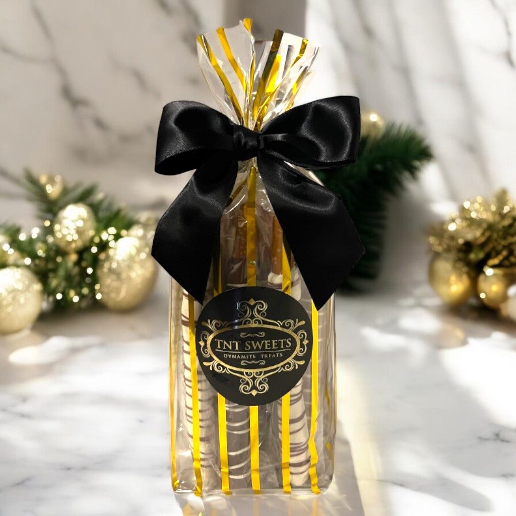 White chocolate, milk chocolate and dark chocolate pretzels in a gold striped bag on a marble countertop with a blurred holiday background.