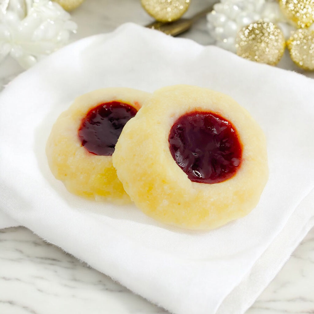 Two Raspberry Thumbprint cookies on a linen napkin on a marble countertop with blurred white and gold decor in the background.