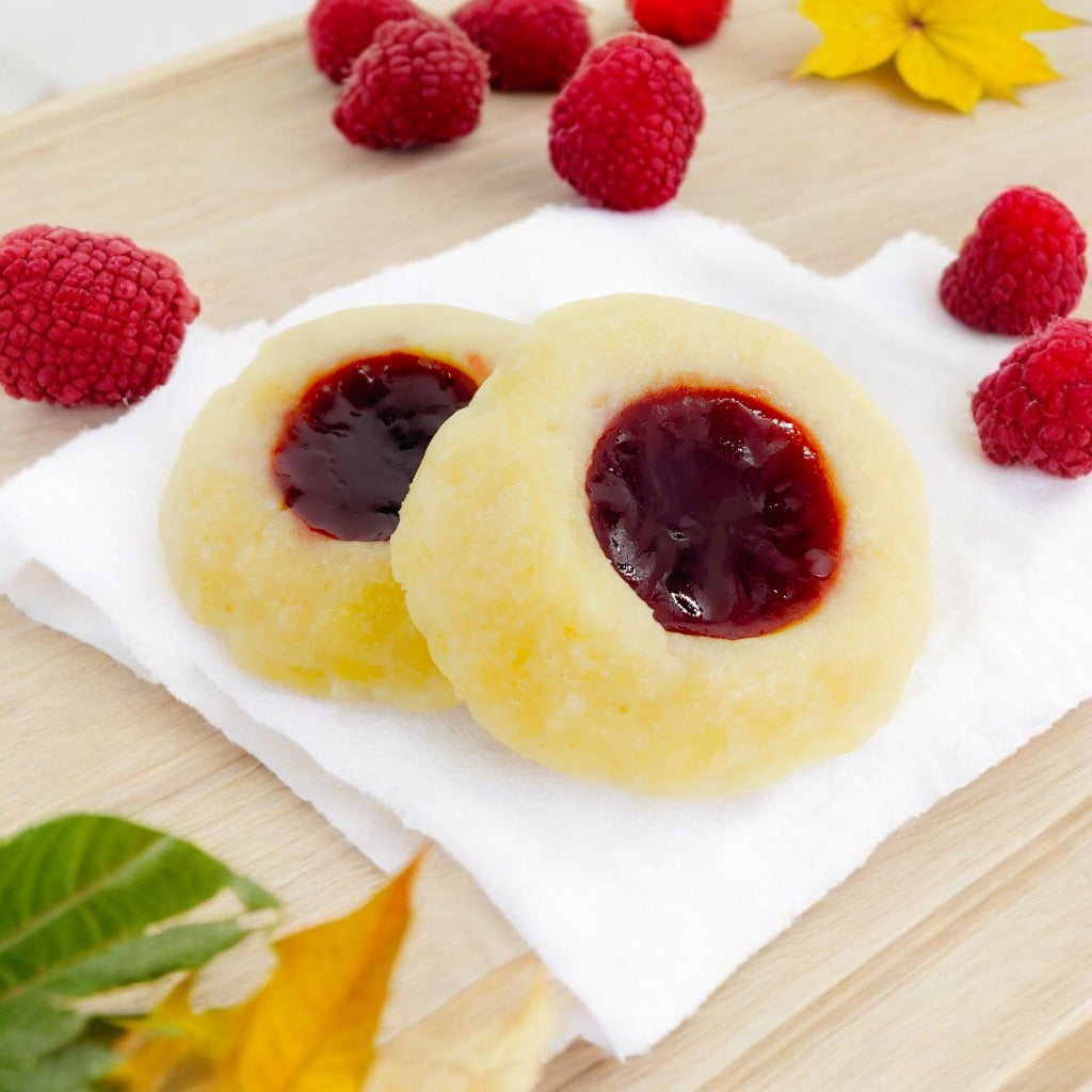 Two Raspberry Thumbprint cookies on a linen napkin with scattered raspberries and scattered fall leaves.