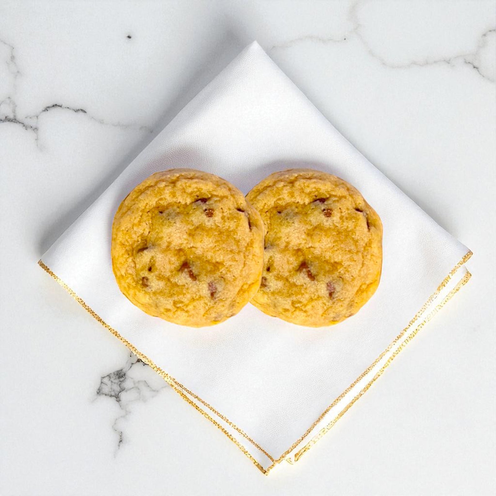 Two signature chocolate chip cookies on a white napkin with gold trim on a marble surface.