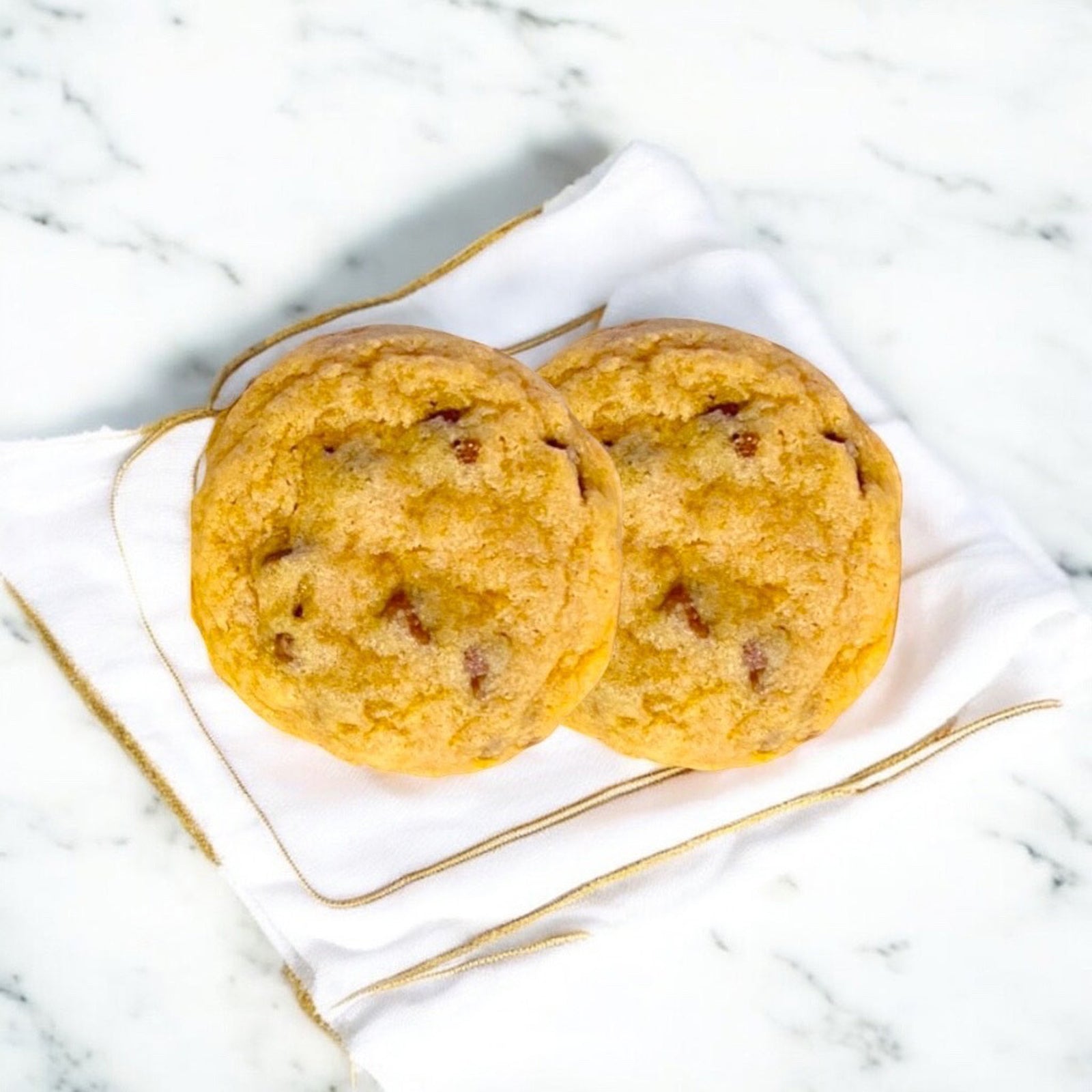 Two chocolate chip cookies on a linen napkin with gold trim on a marble countertop.