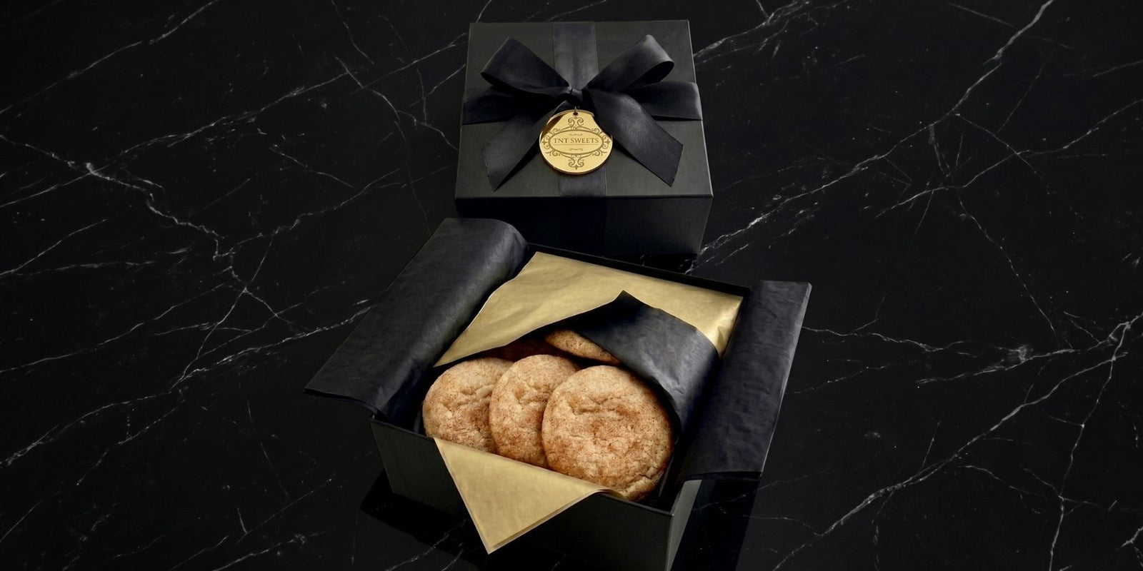 TNT Sweets The Signature cookie assortment featuring twelve gourmet cookies across two signature flavors, presented in a Signature Wrap gift bag with a black satin bow.
