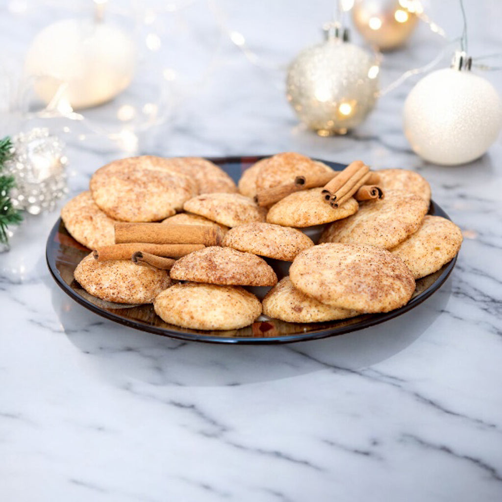 Plate of snickerdoodle cookies with cinnamon sticks on a marble countertop with holiday decor in the background.