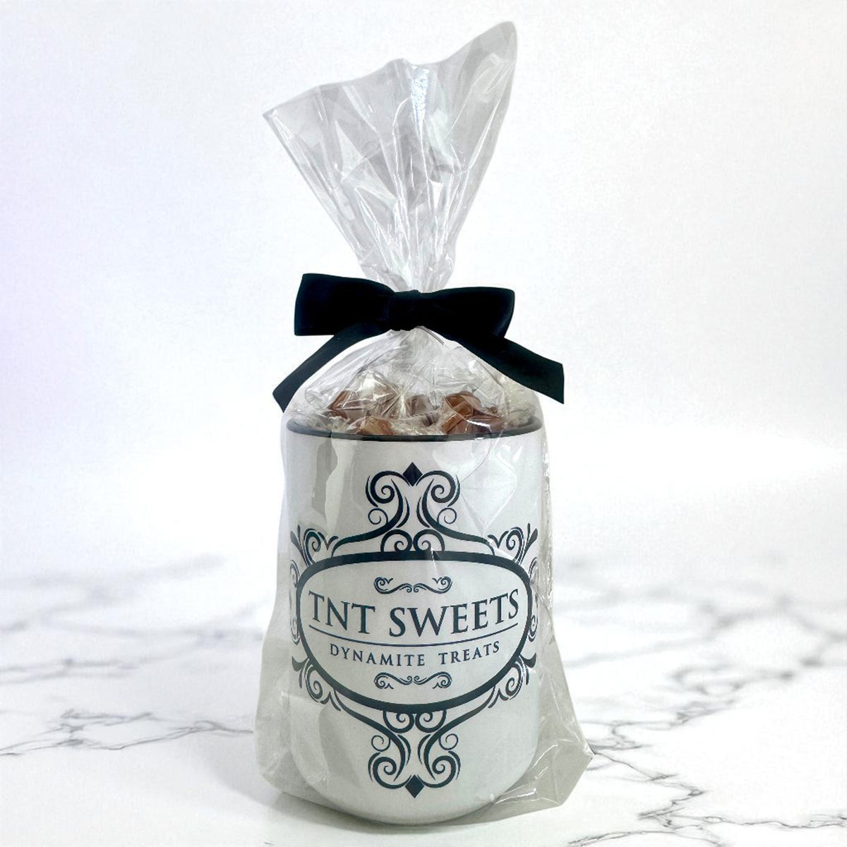15 oz TNT Signature White Mug filled with caramels wrapped in cellophane with a black bow and black logo on a white marble background.