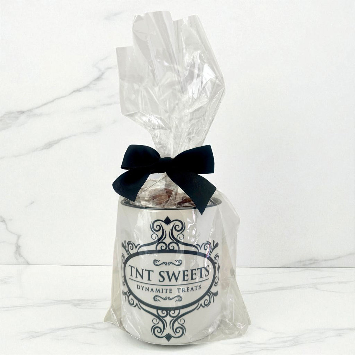 11 oz TNT Signature White Mug filled with caramels wrapped in cellophane with a black bow and black logo on a white marble background.