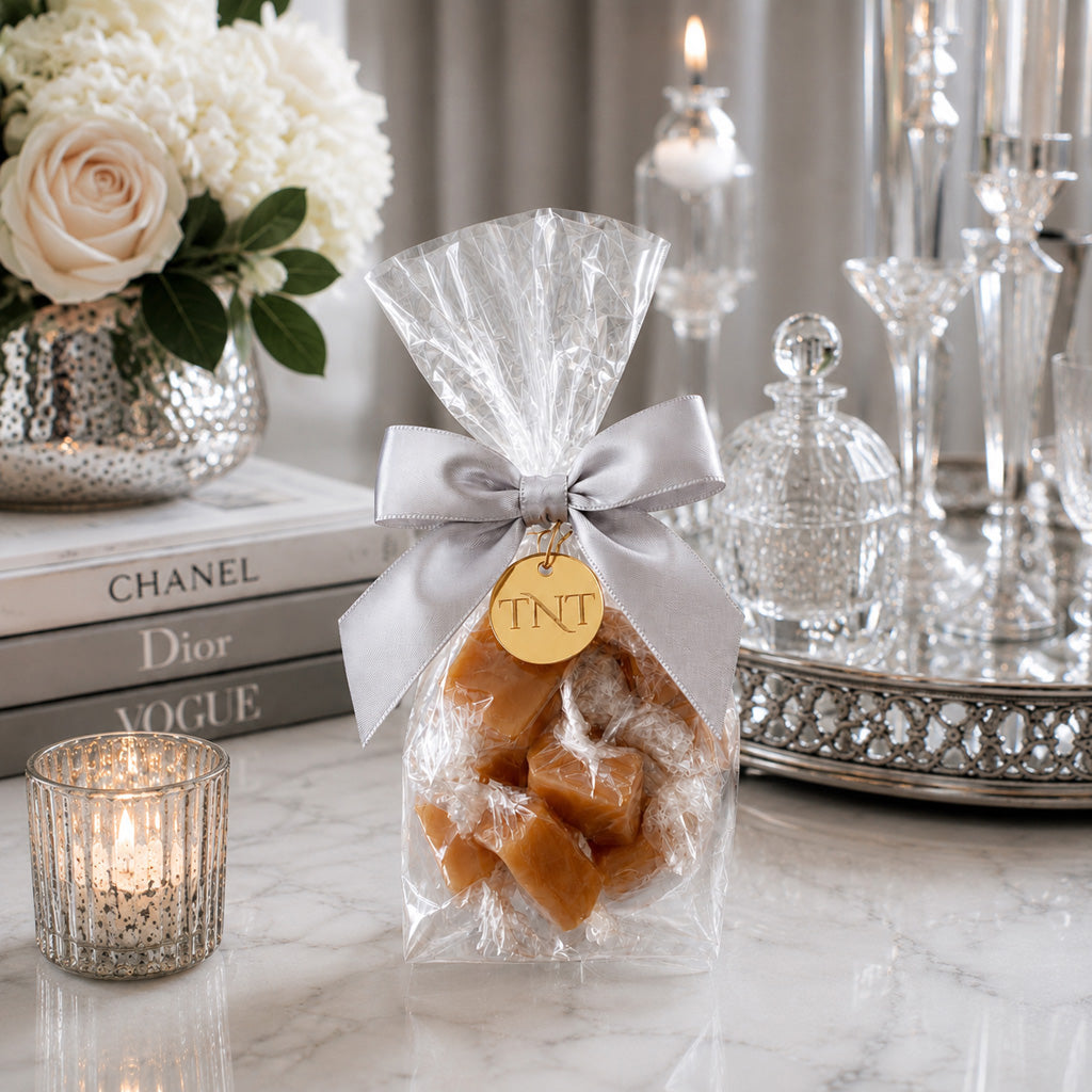 TNT Sweets Amber Velvet caramel favor with silver satin ribbon and gold medallion styled on an elegant silver and white marble table.