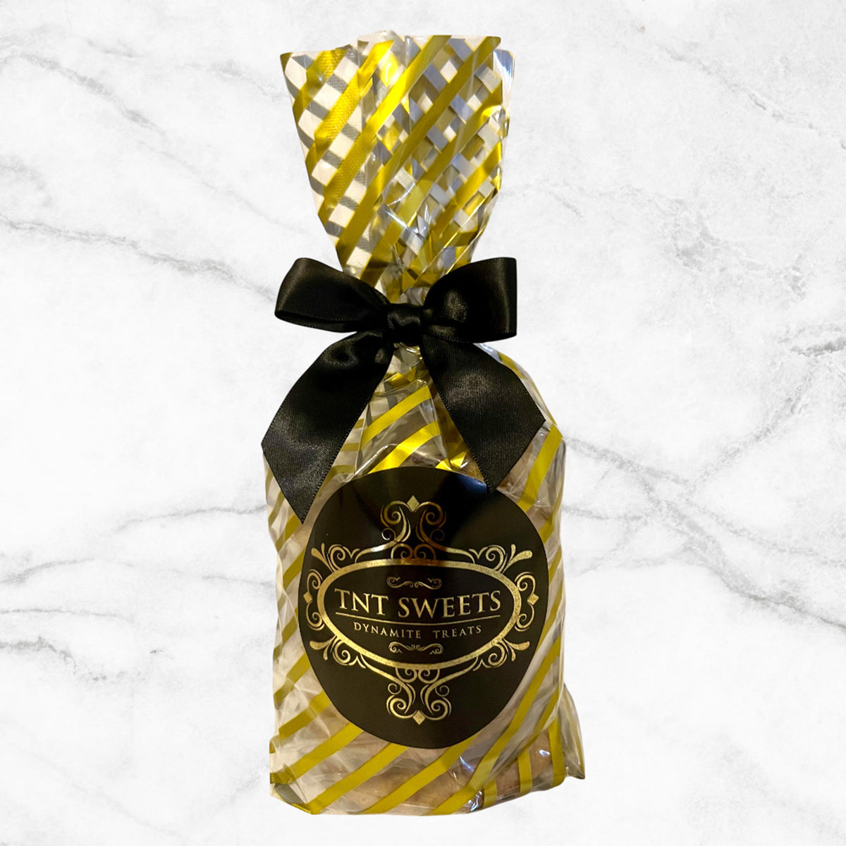 Six cookies in a gold striped cello with a black and gold logo and tied with a black bow on a white marble background.