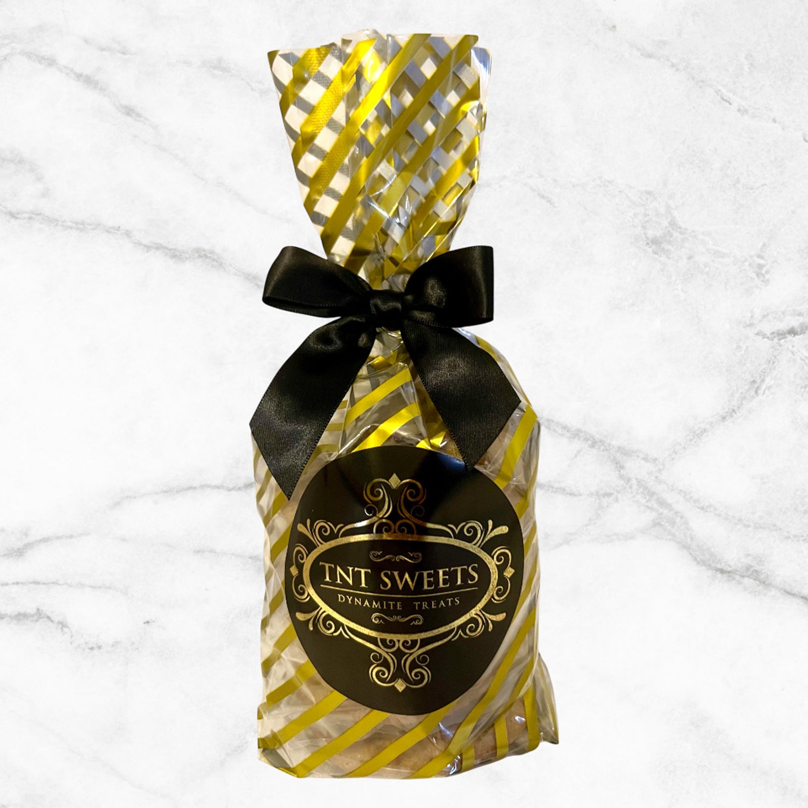Six cookies in a gold striped cello with a black and gold logo and tied with a black bow on a white marble background.
