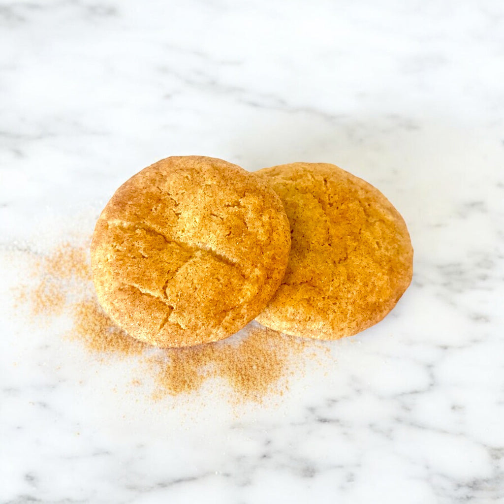 Two Copper Spice Snickerdoodle Cookies from TNT Sweets made with Ceylon cinnamon and Madagascar vanilla, displayed on a white marble surface dusted with cinnamon. 