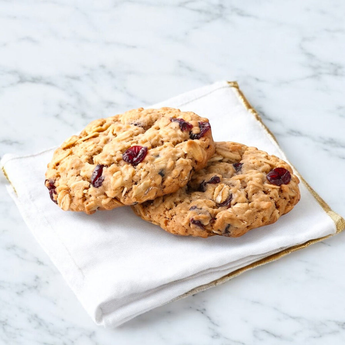 Two Crimson Oat cranberry oatmeal cookies by TNT Sweets made with organic cranberries and premium European butter on a white linen napkin trimmed in gold on a white marble surface.