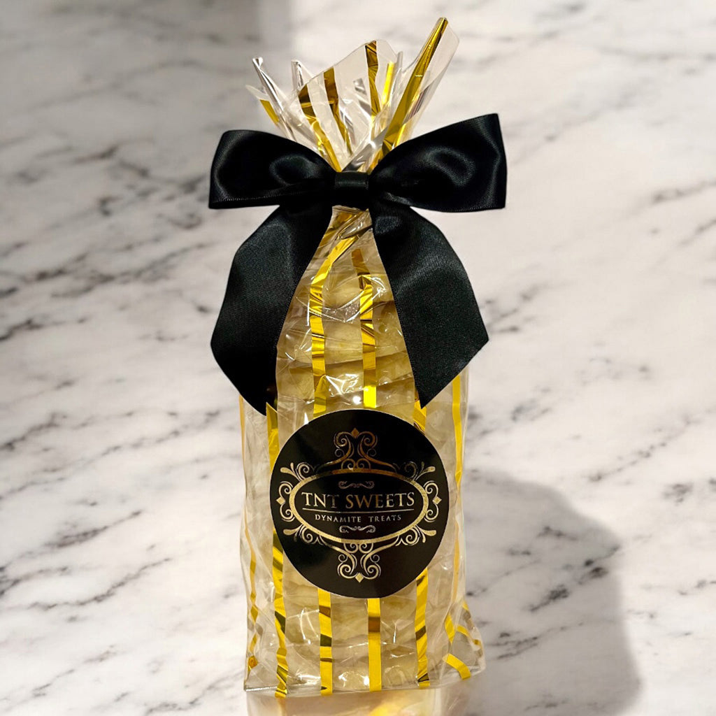 A dozen Lumiere glazed lemon cookies from TNT Sweets individually wrapped, presented in the Gold Stripe Wrap with a black satin bow and Signature TNT Sweets seal, displayed on a white marble surface.