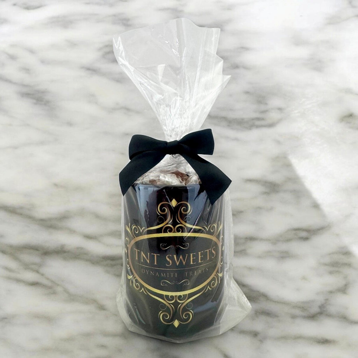 15oz black gift mug filled with caramels wrapped in cellophane with a black bow and a gold TNT logo on a white marble background.