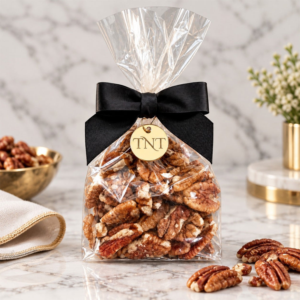 TNT Sweets half-pound pecans in a clear bag with a black satin bow finished with a TNT gold monogram with scattered pecans on a white marble background.