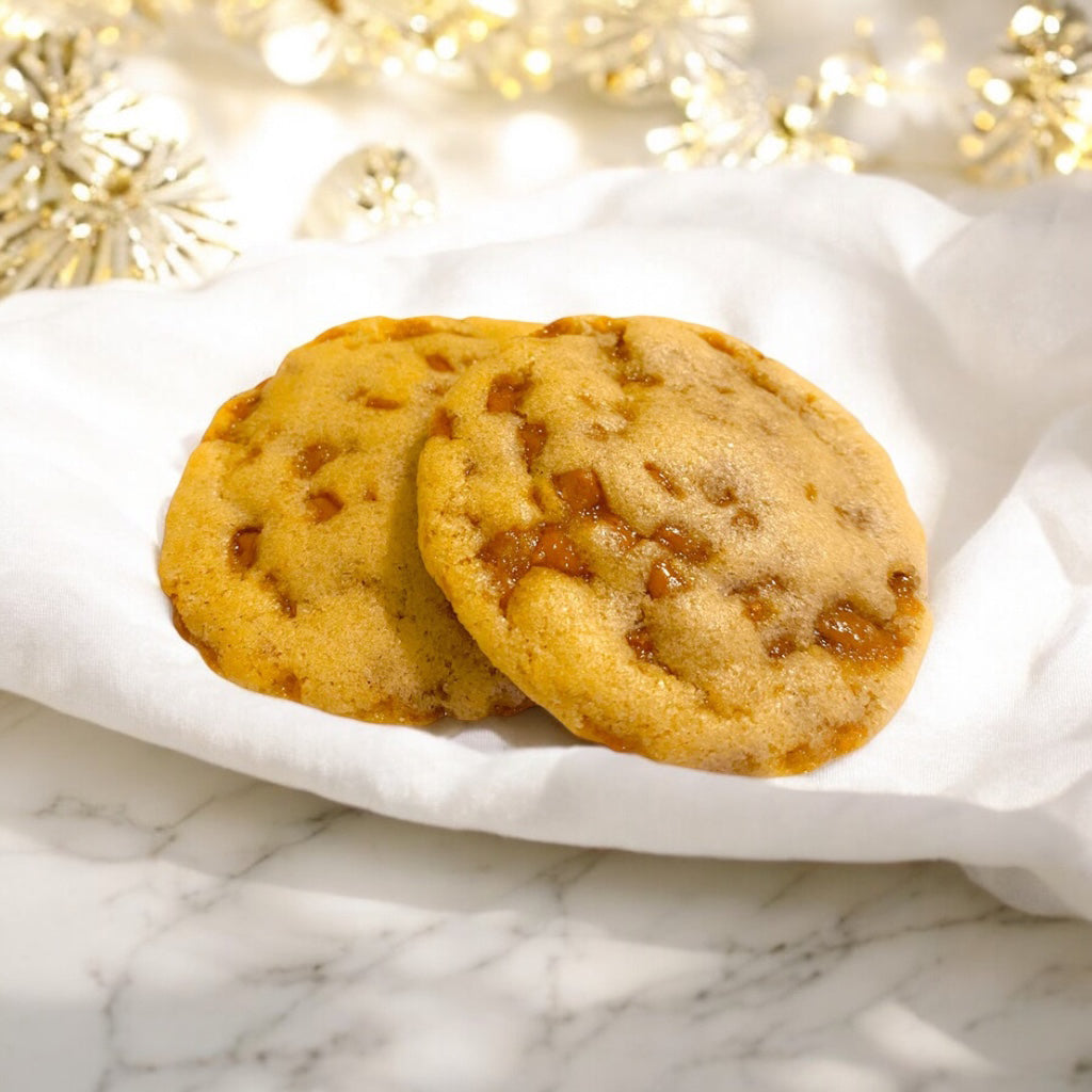 Two Toffee cookies on a linen napkin on a white marble countertop with scattered white decor blurred in the background.