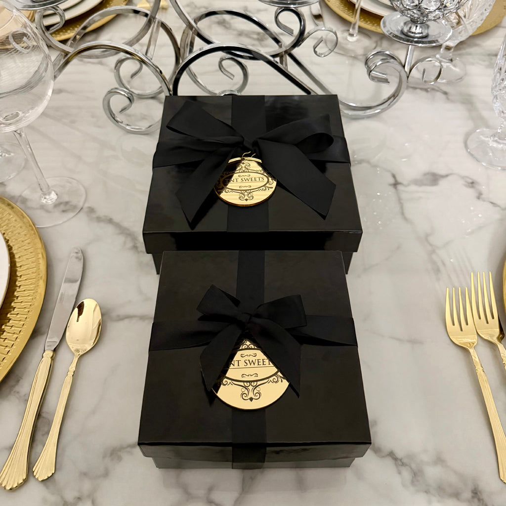 Two Keepsake black gift boxes from TNT Sweets with black satin ribbons and gold acrylic medallions on a white marble dining table with gold place settings.