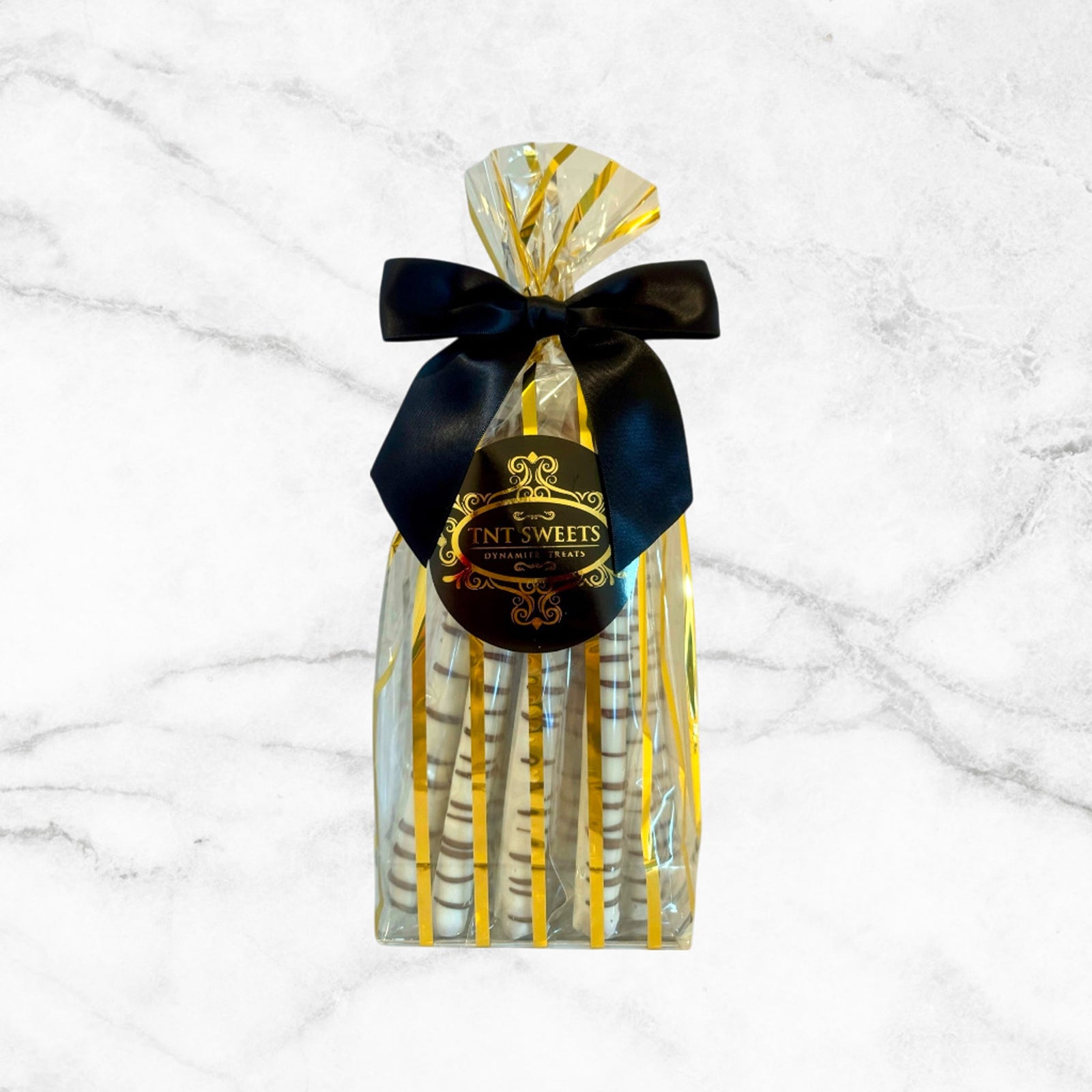 White chocolate dipped pretzels in a gold striped bag with a black bow and label on a marble countertop.